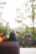 Midnight Mushroom Enamel Mug 16oz next to a kettle on a grill outdoors with trees in the background.