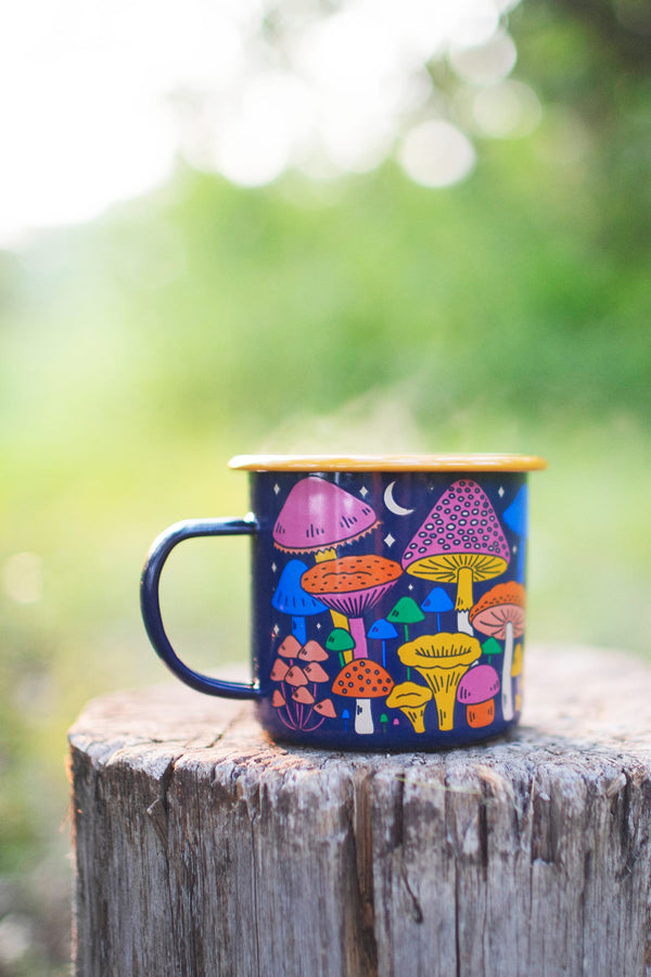 Midnight Mushroom Enamel Mug 16oz with colorful fungi design, resting on a wooden stump. Trek Light, perfect for outdoor adventures.