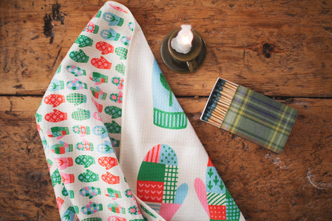 Mittens Kitchen & Tea Towel featuring colorful mitten designs on a wood table with a candle and matchbox.