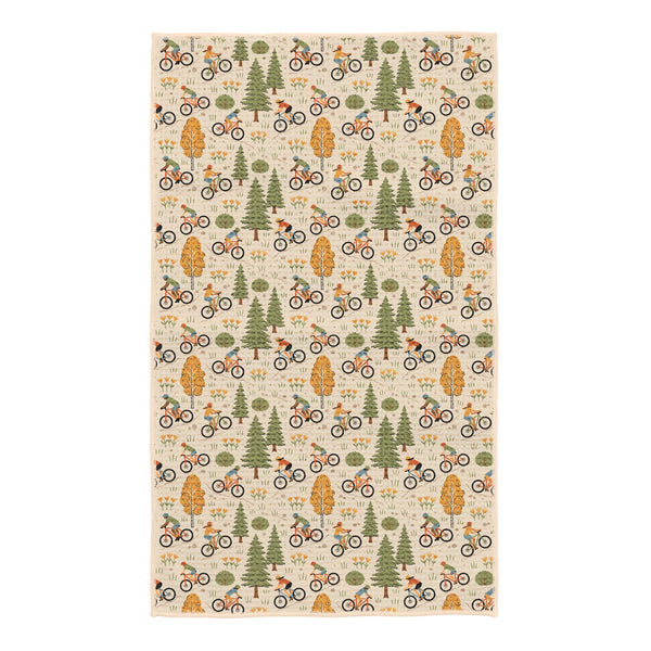 Beige kitchen towel with illustrated mountain bikers riding among green pine trees, aspen trees, and wildflowers