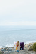 Person holding a cozy Mountain Sunrise Blanket, handwoven from recycled clothing, by the ocean. Beautiful and versatile. By Trek Light.