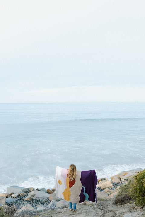 Person holding a cozy Mountain Sunrise Blanket, handwoven from recycled clothing, by the ocean. Beautiful and versatile. By Trek Light.