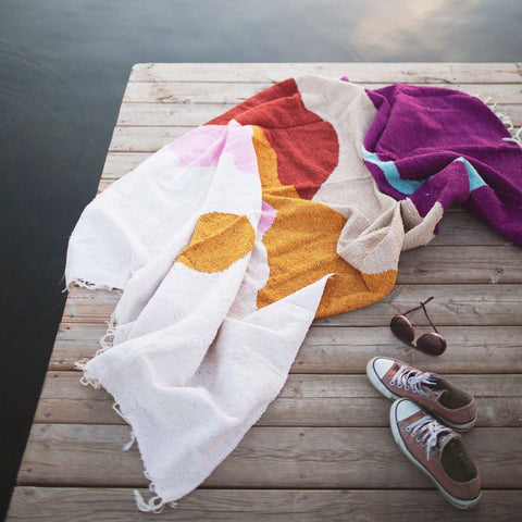 Beautiful handwoven Mountain Sunrise blanket made from recycled clothing materials, cozy and versatile, on a dock beside sneakers. By Trek Light.