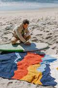 Man on beach admiring colorful Trek Light Mountains Blanket near surfboard, handwoven from recycled materials. Cozy and eco-friendly.