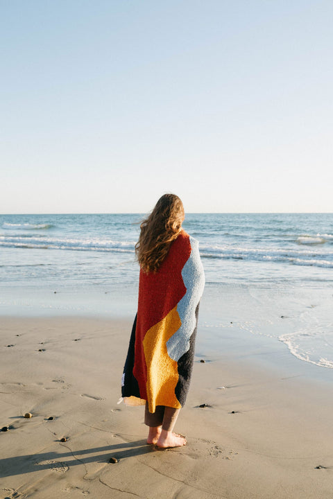 Woman wrapped in colorful Trek Light Mountains Blanket standing on sandy beach facing ocean waves.