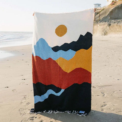 Colorful Trek Light mountain pattern blanket displayed on a sandy beach with ocean waves in the background.