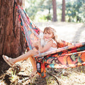 Girl sitting in a colorful hammock, hugging a stuffed animal, enjoying the outdoors with the Double Hammock & Pro Hanging Straps (Midnight Mushroom Print).