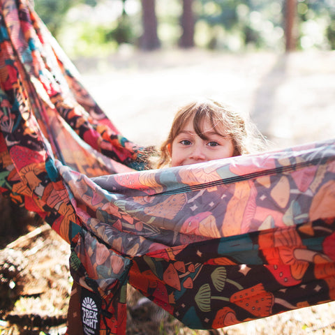 A child peeking out from a colorful Double Hammock & Pro Hanging Straps (Midnight Mushroom Print) in a forest.