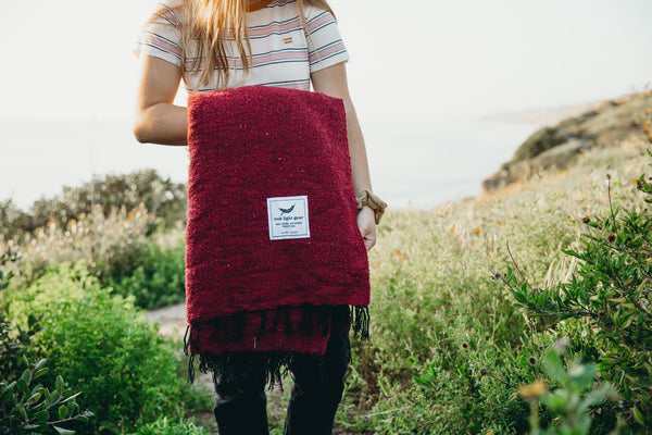 Person holding a red Trek Light Napa Blanket outdoors, made from recycled materials. Eco-friendly and cozy for naps and wine nights.