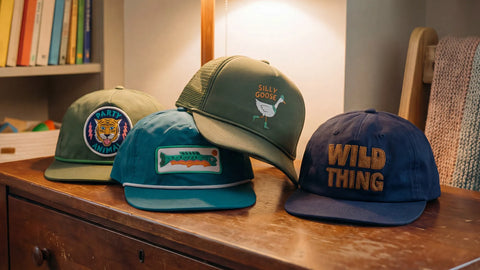 Four trendy caps with unique embroidered designs placed on a wooden dresser, featuring slogans and colorful graphics.