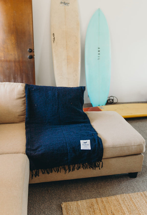 Navy Newport Blanket from Trek Light draped over a beige couch, with surfboards in the background, embodies coastal comfort.