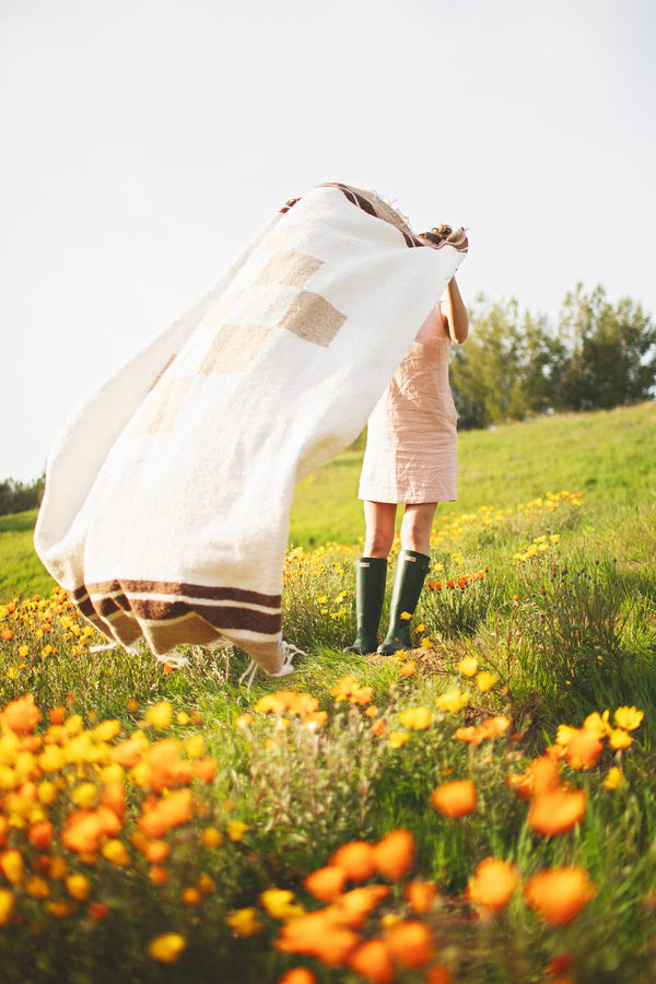 Person joyfully holding a cozy, handwoven North Star Blanket made from recycled clothing materials in a vibrant flower field. By Trek Light.