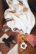 The handwoven North Star Blanket in cream and tan draped over a leather sofa above a rug and boots, Trek Light