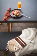 The handwoven North Star Blanket in cream and brown folded over a patio bench beside a breakfast plate, Trek Light