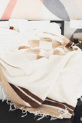 The handwoven North Star Blanket in cream and tan draped across a made bed with white bedding, Trek Light