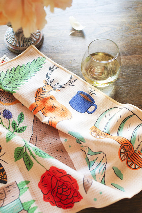 Oregon Kitchen & Tea Towel featuring vibrant designs with a deer and coffee cup on a rustic table.
