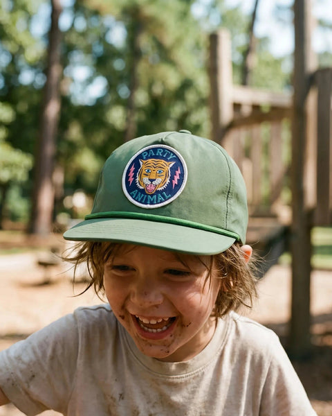 Smiling muddy-faced child wears the green Party Animal Kids Hat with a round tiger patch at a sunny wooded playground.