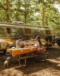 Couple enjoying a picnic on a Peace Blanket outside their Airstream in nature, surrounded by trees.