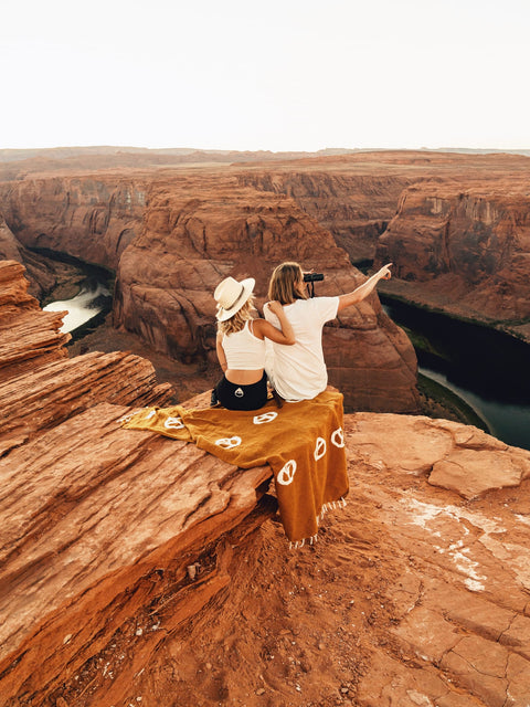 Couple sitting on cliff with a Trek Light Peace Blanket overlooking a canyon, enjoying a scenic view and pointing at the horizon.