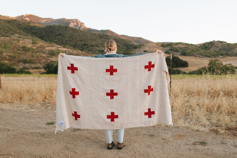 Person holding Trek Light Positive Vibrations Blanket, handwoven with recycled materials, featuring red crosses and a sustainable design.