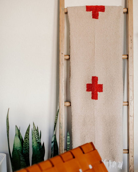 The handwoven Positive Vibrations Blanket in cream with red crosses hung on a wooden ladder beside a snake plant, Trek Light