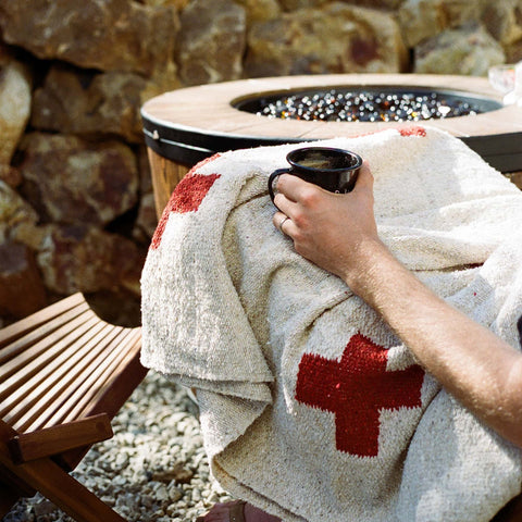 Person relaxing by a stone fire pit, wrapped in a cozy wool blanket with red cross embroidery, holding a black mug.