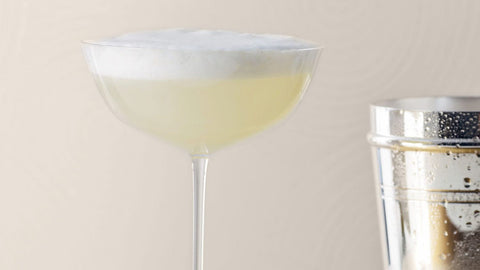 Elegant cocktail with frothy top in a stylish glass beside a shaker.