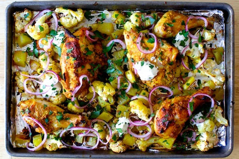 Roasted chicken legs with cauliflower, potatoes, and red onions on a baking tray, garnished with fresh herbs.