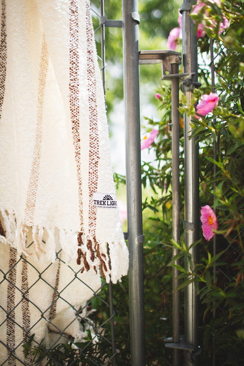 Handwoven Silverton blanket with stripes, made from recycled materials, hanging near pink flowers outdoors.