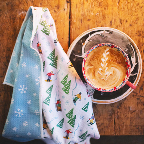 Snow Day Kitchen & Tea Towel with winter design beside a latte on a wooden table. Snowflake design, super absorbent, quick-drying kitchen towel.