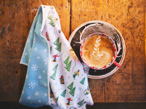 Snow Day Kitchen & Tea Towel with winter design beside a latte on a wooden table. Snowflake design, super absorbent, quick-drying kitchen towel.