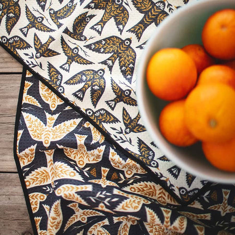 Songbird Kitchen & Tea Towel with bird pattern next to a bowl of oranges on a wooden counter, eco-friendly and super absorbent.