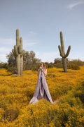Woman wrapped in a lavender handwoven Sonoma Blanket, standing in a cactus-filled desert landscape, exuding beauty and coziness. By Trek Light.
