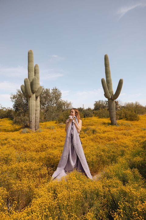 Woman wrapped in a lavender handwoven Sonoma Blanket, standing in a cactus-filled desert landscape, exuding beauty and coziness. By Trek Light.