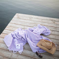 Lavender Sonoma Blanket, handwoven from recycled materials, on a dock with a straw hat and sunglasses, exuding cozy and versatile charm. By Trek Light.