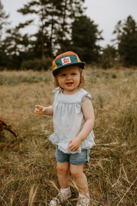 Child wearing colorful Trek Light Stay Wild hat, smiling in a field, ready for outdoor adventure. Perfect 5-panel camper style for kids.