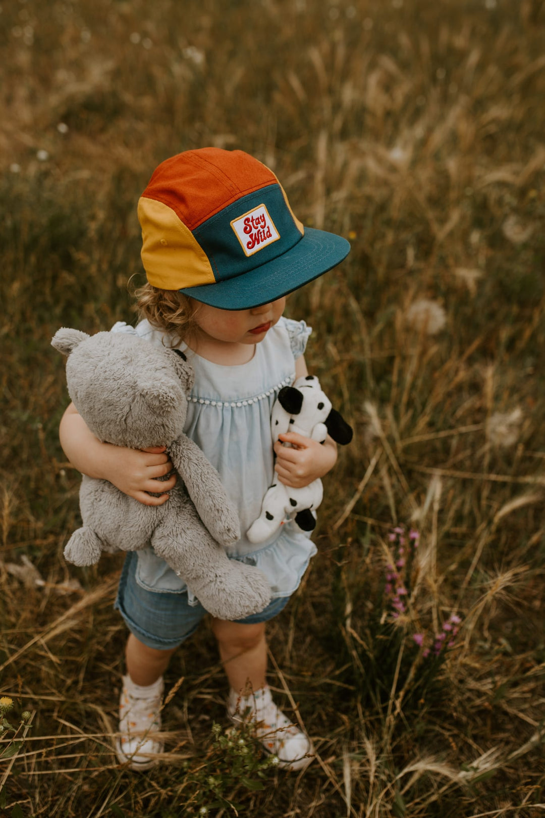 Child wearing the colorful Stay Wild kids hat, holding stuffed animals in a grassy field