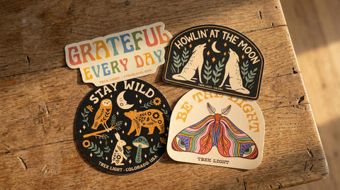 Colorful stickers on wooden table featuring designs with phrases like 'Grateful Every Day' and 'Be the Light' by Trek Light.