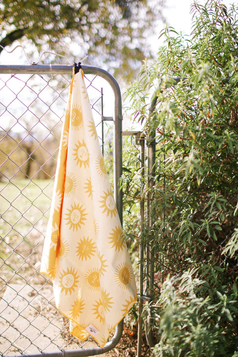 Sunny Wander & Travel Towel hanging on a fence, showcasing its bright sunshine design.