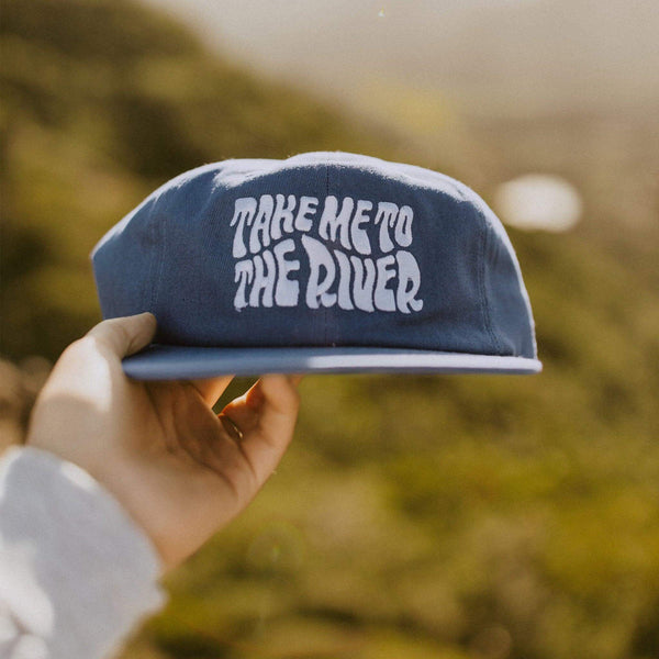 Hand holding a blue 'Take Me To The River' 6-panel snapback hat, perfect for river days, fishing, and adventures with Trek Light.