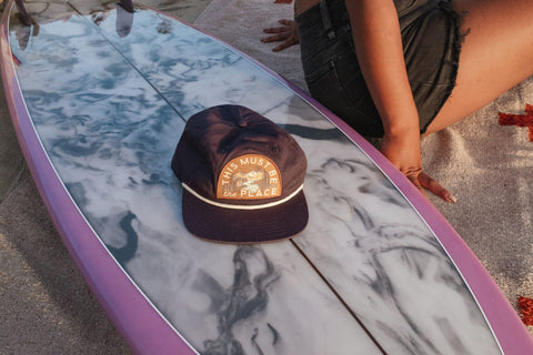 This Must Be The Place Hat (Mountains) placed on a surfboard, perfect for beach adventures.
