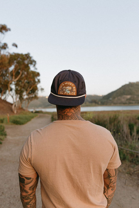 Person wearing This Must Be The Place Hat (Mountains) while walking on a scenic pathway near water.