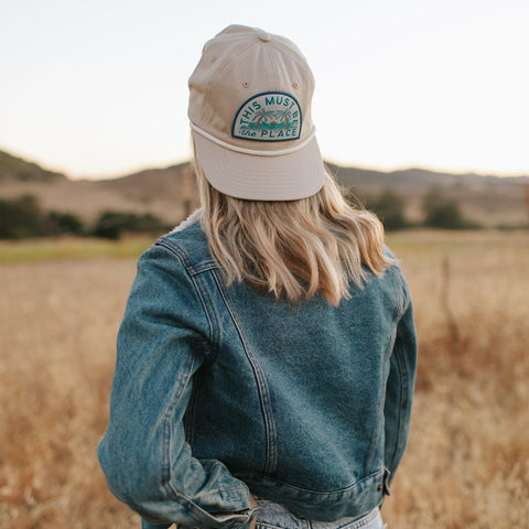 Woman in denim jacket wearing a beige 'This Must Be The Place' hat by Trek Light, enjoying scenic outdoor view.