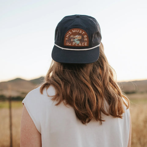 This Must Be The Place Hat (Mountains) worn by a person with long hair in a scenic outdoor setting.