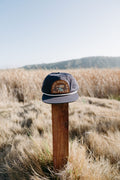 This Must Be The Place Hat (Mountains) displayed on a wooden post in a grassy field with mountains in the background.