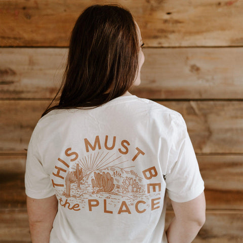 Person wearing a "This Must Be The Place" shirt, handprinted in the USA with Trek Light design, showcasing desert-themed illustration.