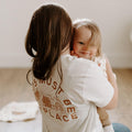 Mom holding child wearing 'This Must Be The Place' shirt by Trek Light, showcasing super soft cotton feel and handprinted design.