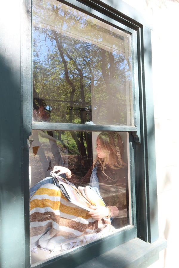 Woman reading while wrapped in Golden Age Blanket by a window, enjoying comfort and the view outside.