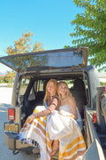 Two women wrapped in Golden Age Blankets sitting in the back of a vehicle, enjoying a sunny day outdoors.