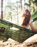 Girl with a braid relaxes in a green Double Hammock (Trout) with fish and star print in a sunny forest.